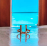 A minimalist rose gold ring with a double-bar design, resting on a reflective blue surface with a blurred window and warm wooden frame in the background.