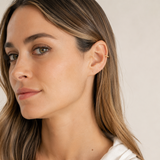 A close-up of a woman with long brown hair wearing a delicate gold ear cuff on her ear lobe, set against a soft beige background.