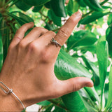 A silver ring with a small green gemstone is worn on a person's finger, shown against a background of lush green leaves. The person also wears a thin silver wire bracelet on their wrist.