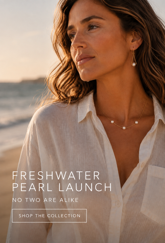 A woman on a beach at sunset wearing delicate pearl jewelry, including a necklace and earrings, with a white button-up shirt. Text overlay reads 'FRESHWATER PEARL LAUNCH' and 'NO TWO ARE ALIKE' with 'SHOP THE COLLECTION' below.