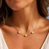 A close-up of a woman wearing a delicate gold necklace with three small white pearls spaced along the chain, styled with a white V-neck top.