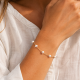 A delicate gold chain bracelet with seven small white pearls, worn on a person's wrist. The person is wearing a white linen top, and the background is softly blurred.