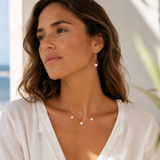 A woman wearing a delicate gold necklace with three small white pearls and matching drop earrings, styled with a white linen top against a bright, sunlit background.