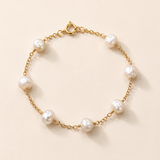 A delicate gold chain bracelet featuring seven irregular white freshwater pearls spaced evenly along the chain, displayed on a warm beige background.